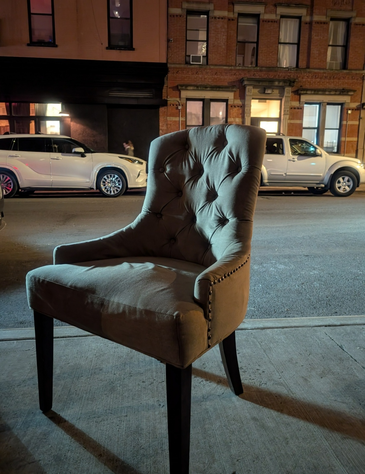 a regal chair sits near a curb in the night, faced away from the street, side-lit by a vehicle or something