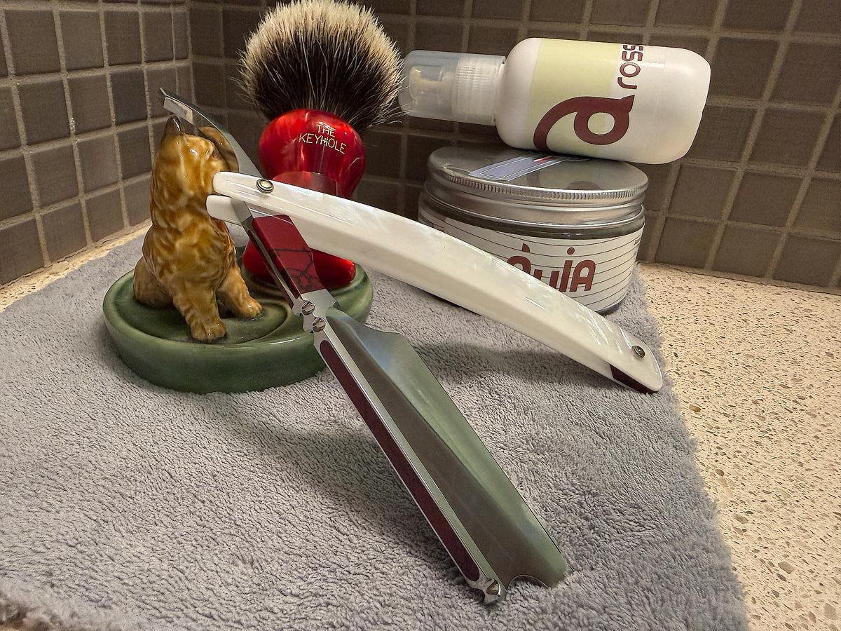 On a gray microfiber towel a straight razor with blade open about 45 degrees leans next to a bottle of aftershave and a jar of shave soap. A shave brush rests bristles-up in a green Wade ceramic pipe rest that features a brown Cairn Terrier.