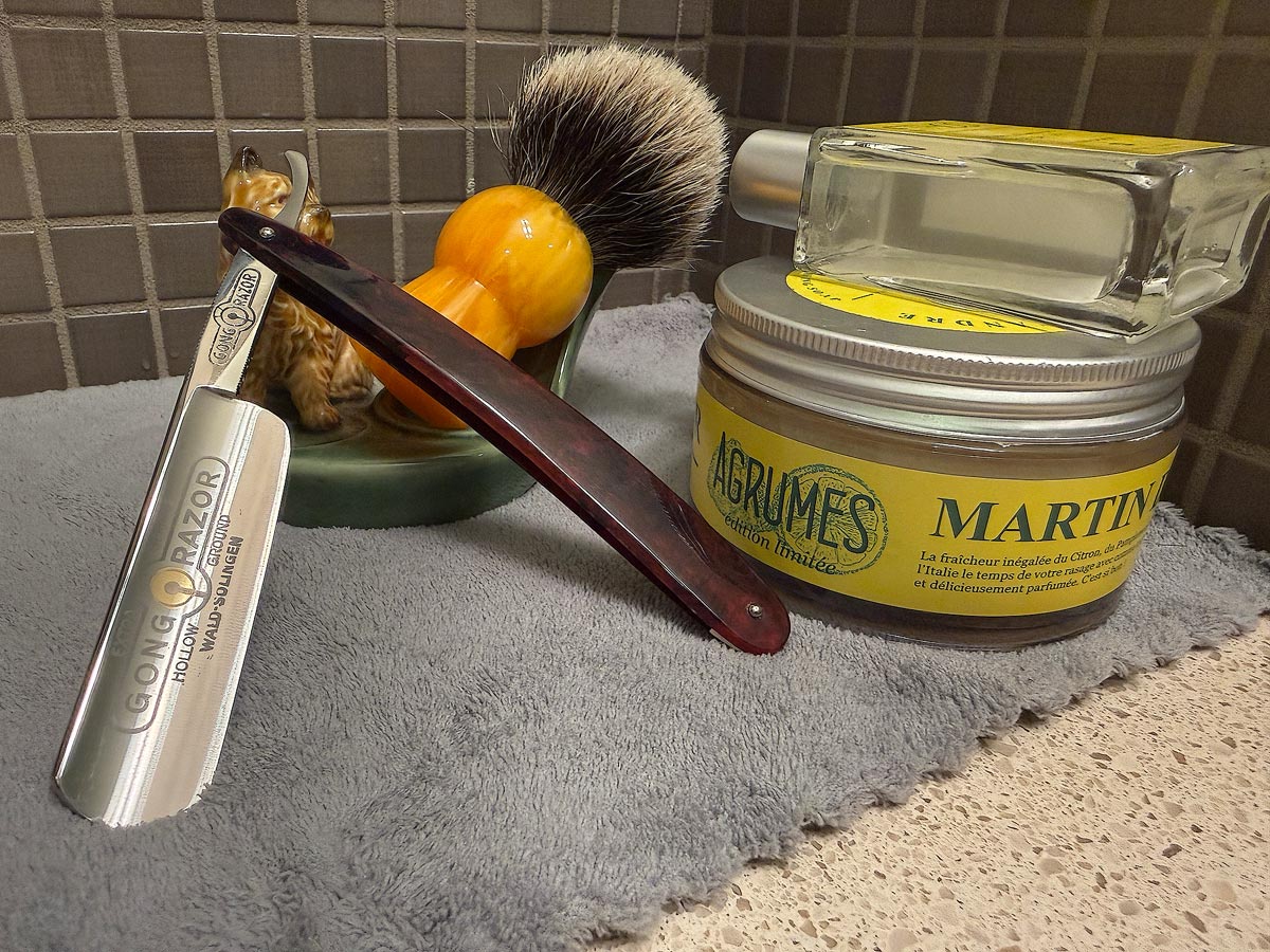On a gray microfiber towel a straight razor with blade open about 45 degrees leans next to a bottle of aftershave and a jar of shave soap. A shave brush rests bristles-up in a green Wade ceramic pipe rest that features a brown Cairn Terrier.