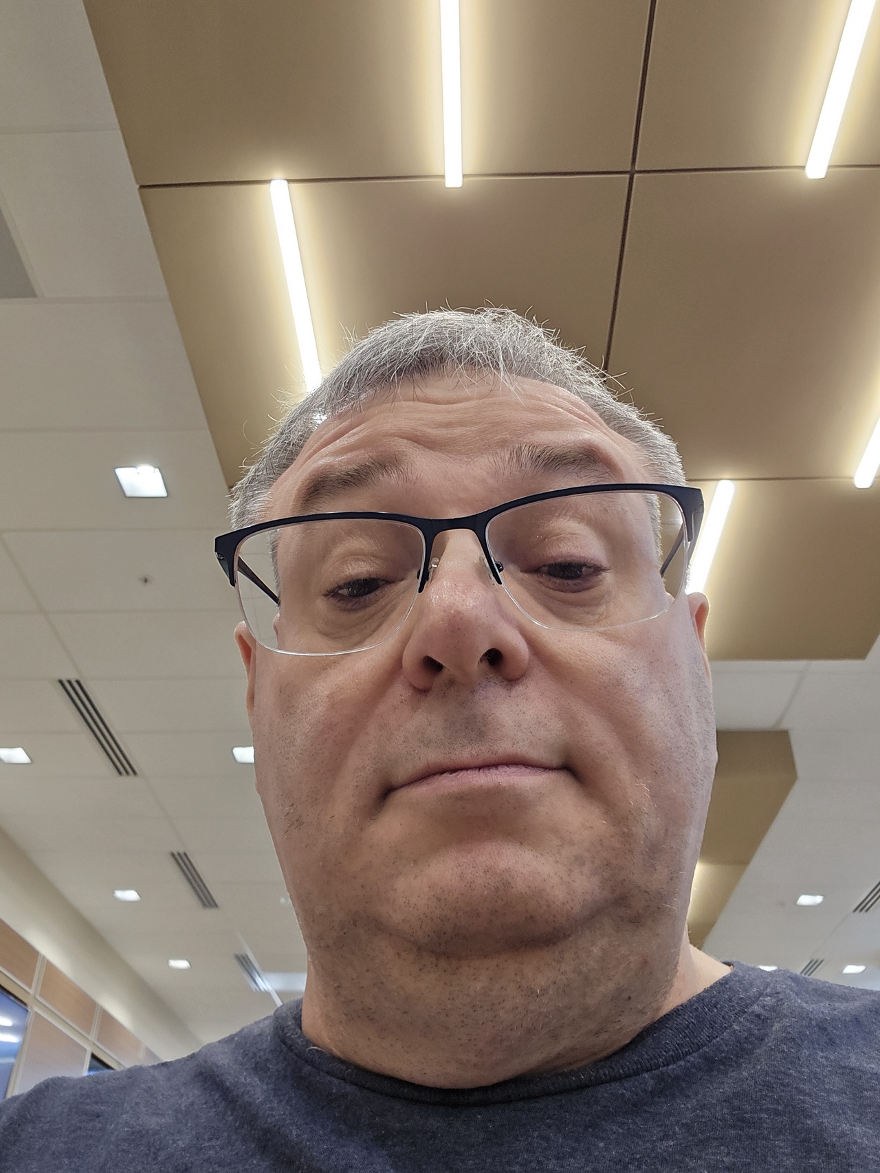 Close-up selfie of a middle-aged man with short gray hair and light skin, wearing rectangular black-framed glasses. The photo is taken from a low angle, capturing the lower half of his face prominently, including his nose, mouth, and chin, with a neutral expression. He is wearing a dark blue shirt. The background shows an indoor setting with a ceiling featuring beige panels and long, bright fluorescent lights arranged in parallel lines. Additional ceiling elements include white square tiles and air vents. The lighting is bright and evenly distributed.