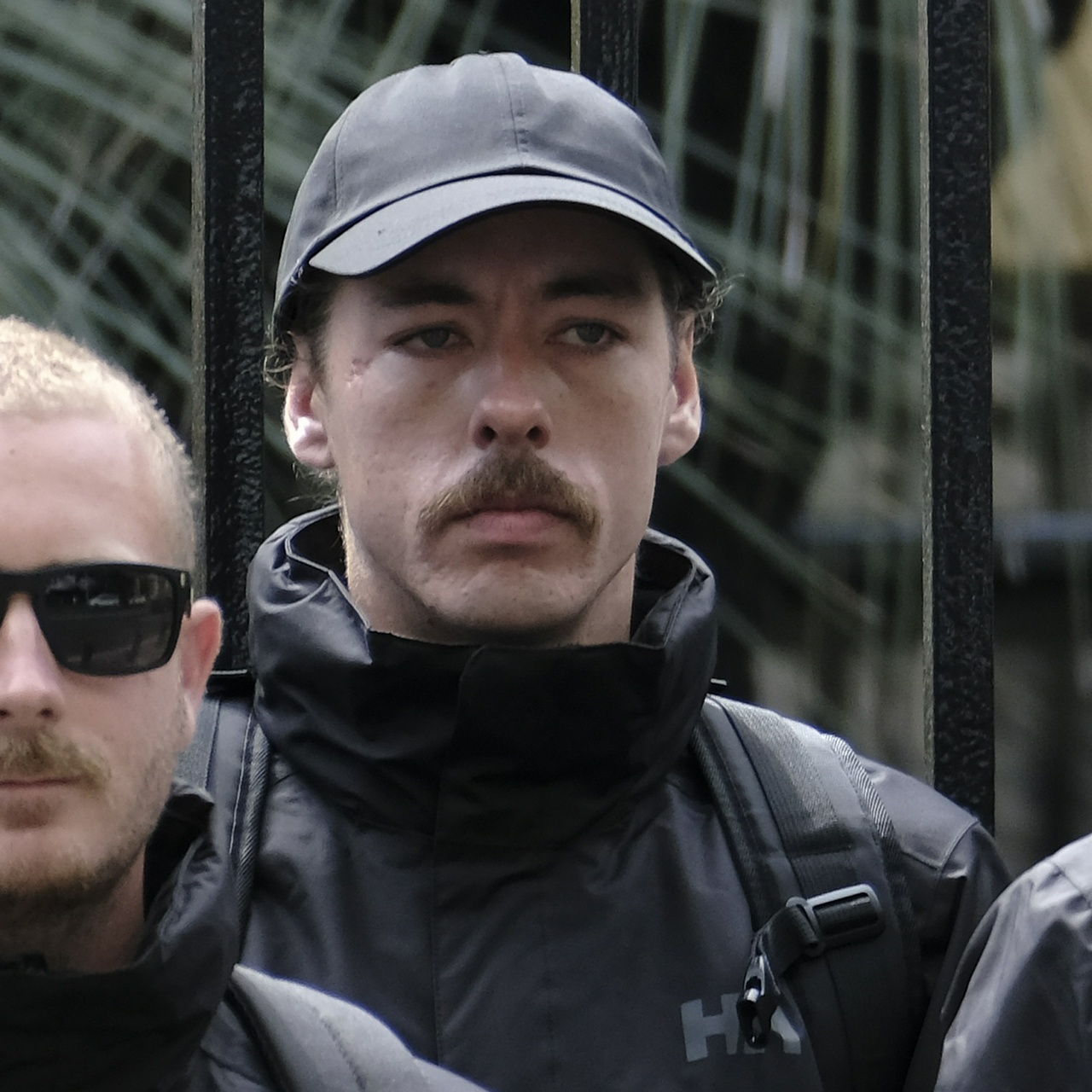 A taller skinny man with a boxy jaw, melancholy eyes and a walrus moustache with large ears tucked into his cap