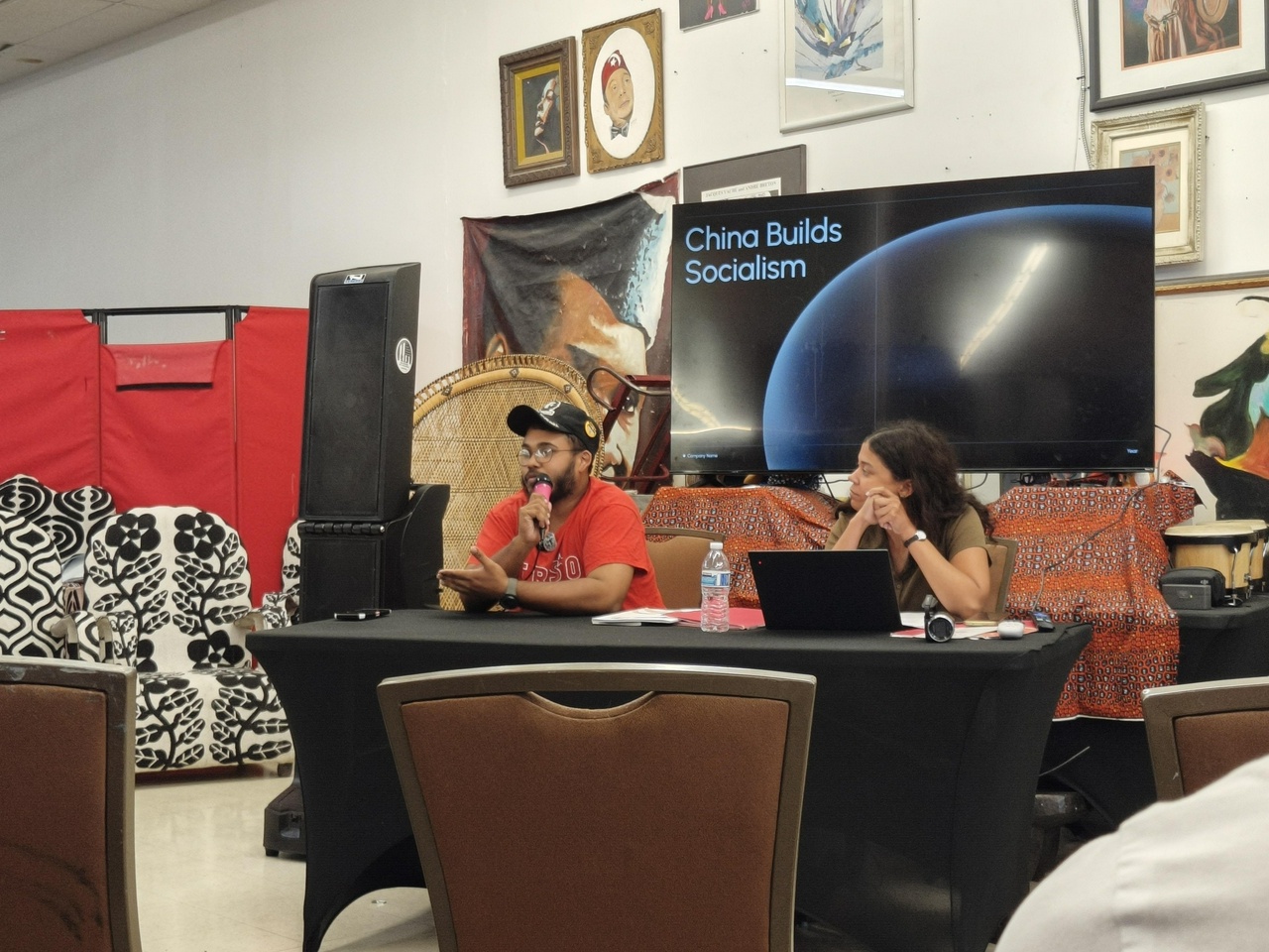Two panelists sit at a table wearing FRSO shirts and giving talks on socialist construction in China.