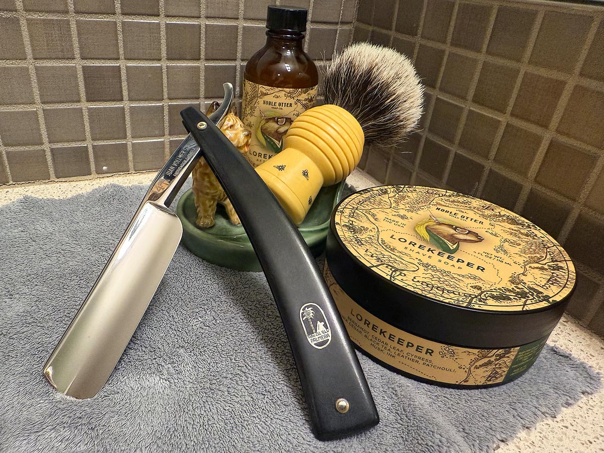 On a gray microfiber towel a straight razor with blade open about 45 degrees leans next to a bottle of aftershave and a jar of shave soap. A shave brush rests bristles-up in a green Wade ceramic pipe rest that features a brown Cairn Terrier.