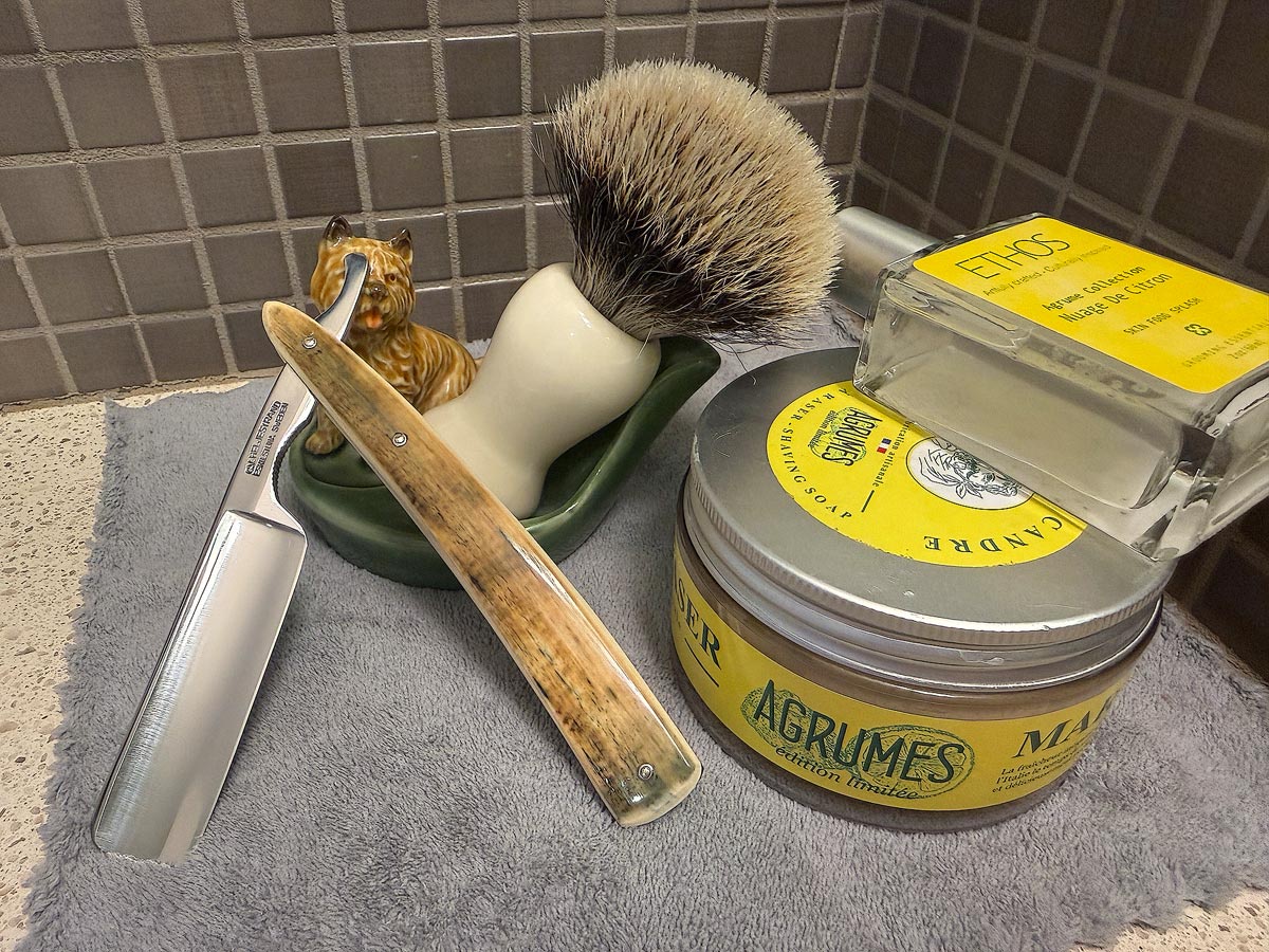 On a gray microfiber towel a straight razor with blade open about 45 degrees leans next to a bottle of aftershave and a jar of shave soap. A shave brush rests bristles-up in a green Wade ceramic pipe rest that features a brown Cairn Terrier.
