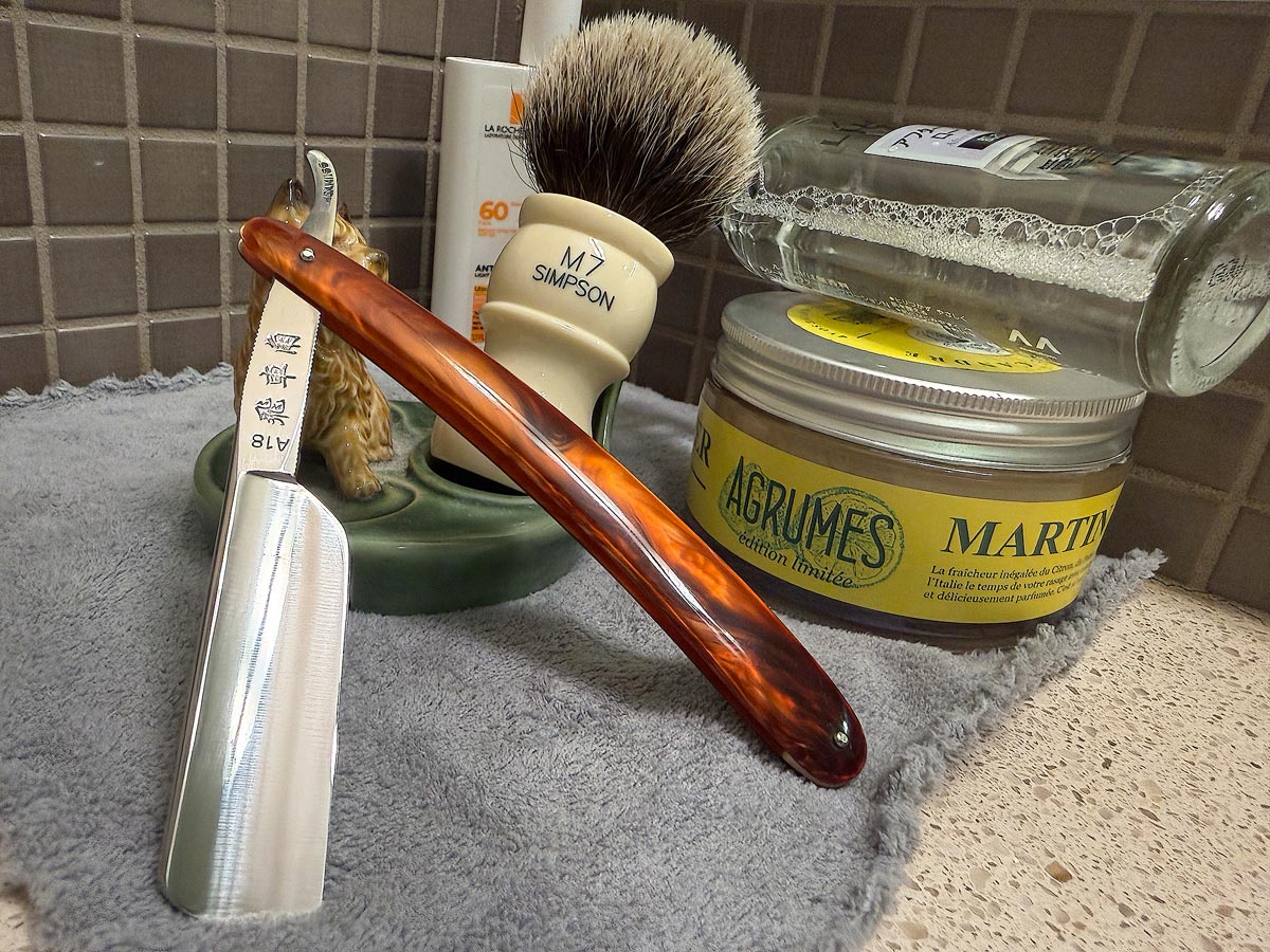 On a gray microfiber towel a straight razor with blade open about 45 degrees leans next to a bottle of aftershave and a jar of shave soap. A shave brush rests bristles-up in a green Wade ceramic pipe rest that features a brown Cairn Terrier.