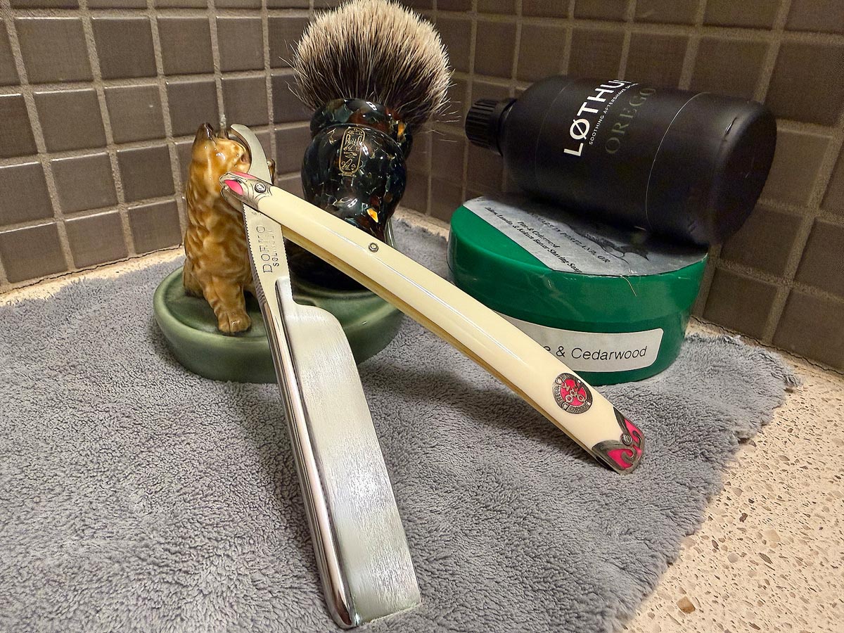 On a gray microfiber towel a straight razor with blade open about 45 degrees leans next to a bottle of aftershave and a jar of shave soap. A shave brush rests bristles-up in a green Wade ceramic pipe rest that features a brown Cairn Terrier.