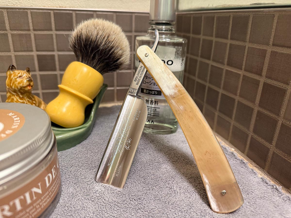 On a gray microfiber towel a straight razor with blade open about 45 degrees leans next to a bottle of aftershave and a jar of shave soap. A shave brush rests bristles-up in a green Wade ceramic pipe rest that features a brown Cairn Terrier.